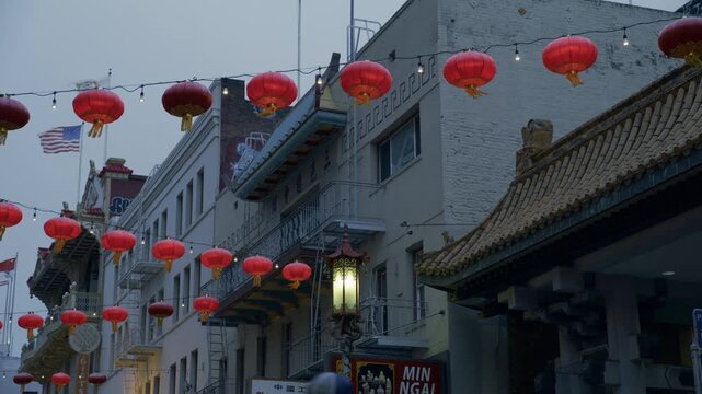 Red Lanterns Strung Over Chinatown Street Buildings And American Flag Caught In Wind - Low Shot