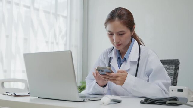 Female doctor using a smartphone while working at a desk with a laptop in a clinic, representing telehealth, digital healthcare, mobile communication, and modern medical technology.
