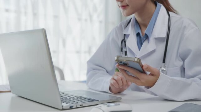 Close-up of a doctor holding and using a smartphone beside a laptop in a medical office, representing telemedicine, telehealth communication, and digital healthcare workflow.