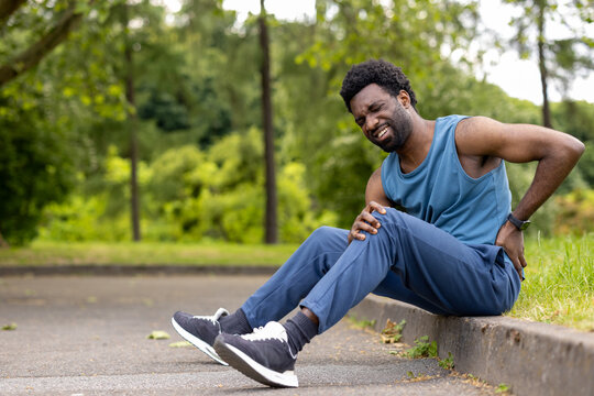 Young man sitting on the curb of an asphalt path, holding his knee and lower back with a grimace of pain, signaling a sports injury while exercising outdoors in a park