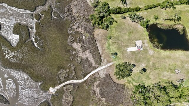 Aerial view of the Tolomato River Boardwalk Preserve with a wooden walkway extending into marshlands, surrounded by green grass, trees, and a pond in St. Augustine, Florida, United States.