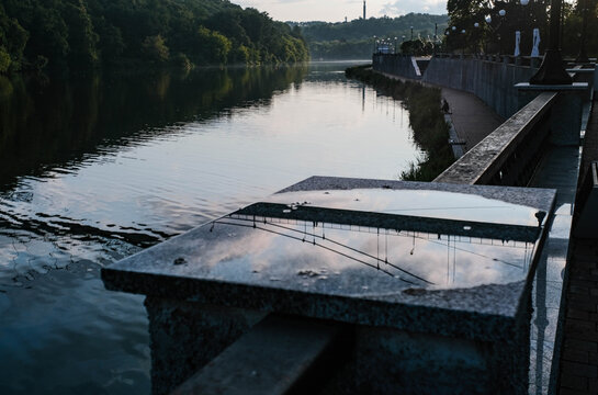 Bridge reflection in puddle on granite pier next to calm river at sunrise