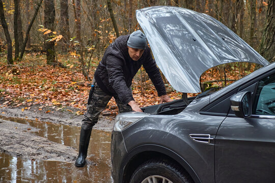 Man inspecting a broken car with open hood on a muddy forest road in autumn. Vehicle breakdown, roadside assistance, travel problem and transportation emergency concept