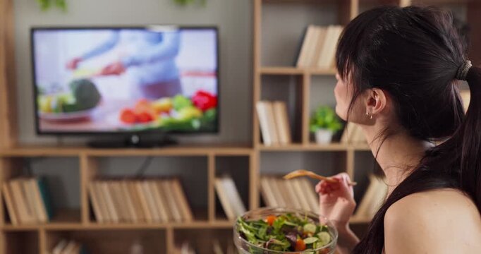 Woman eating salad at home watching tv. She holds a bowl and enjoys fresh greens while a cooking show plays, promoting nutrition and wellness at home. Clear concept of healthy diet habits.