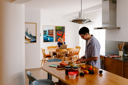 A man chops food while a blonde woman sits at a laptop behind him.