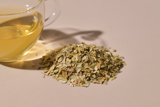 Pile of dried yerba mate leaves next to a glass cup of brewed tea