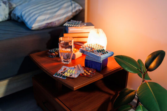 Various blister packs with medicine lay on a bedside table under warm lamp light late at night