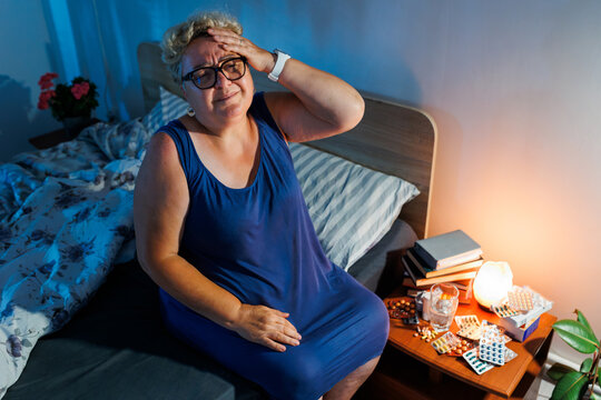 Senior woman experiencing intense headache near medical pills inside bedroom.