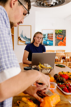 A man chops fresh fruit while a blonde woman works on her laptop.