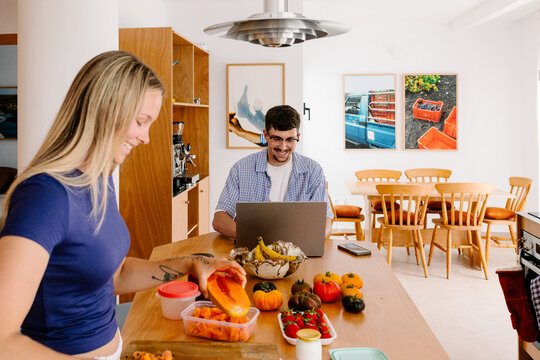 A smiling woman preps fruit while a man works on his laptop.
