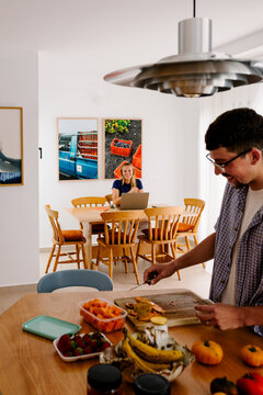 A man preps food while a blonde woman works on a laptop nearby.