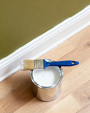 A paint brush resting on a metal tin of white wall paint placed on a wooden floor near a freshly painted green interior wall baseboard