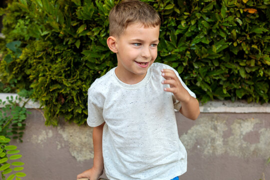 A happy young boy smiles softly while looking to the side, standing outdoors near a lush green hedge in a casual candid moment