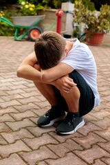 Obraz premium Sad teenage boy sitting on pavement with head on folded arms, expressing loneliness, stress, emotional pain and depression in an outdoor urban setting alone