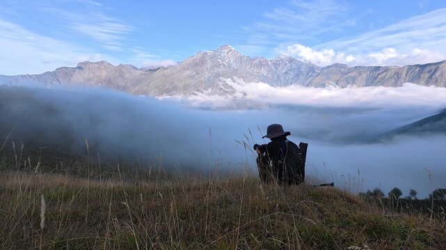 4K real time video of a solo traveler sitting on a grassy mountain hill looking down at thick white fog.
