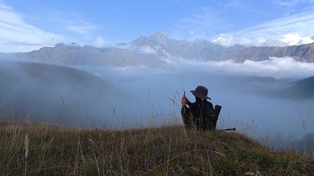 4K real time video of a solo hiker sitting on a grassy hill touching hat while looking at mountains over thick fog.