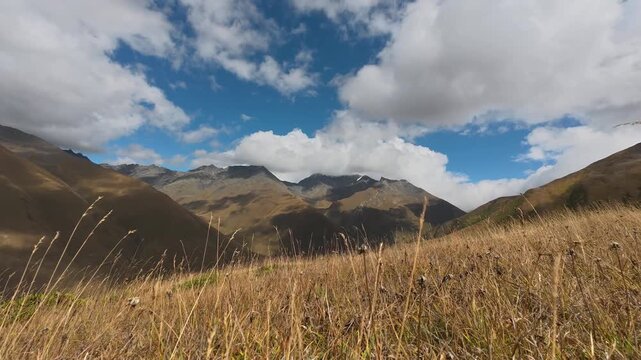 4K timelapse video of white clouds moving across the blue sky casting shadows over wild mountain range and golden grass.