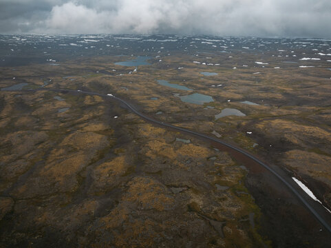 Aerial view of the rugged Westfjords Region landscape in Iceland, featuring a winding road cutting through mossy terrain, scattered small ponds, and patches of snow under a cloudy sky. Iceland.
