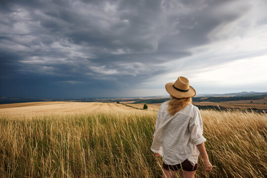 Worried female farmer looking at approaching storm clouds over agricultural field. Summer weather and impact climate change on agriculture concept