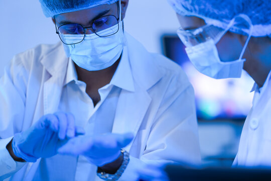 Laboratory colleagues hold a vial up while analysing results in a dramatic blue-lit laboratory while wearing white lab coats and blue protective gloves and modern scientific innovation.