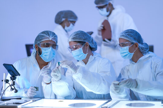 A semiconductor inspection team study a reflective silicon wafer closely in a blue-lit semiconductor cleanroom in protective hairnets, masks and safety goggles.