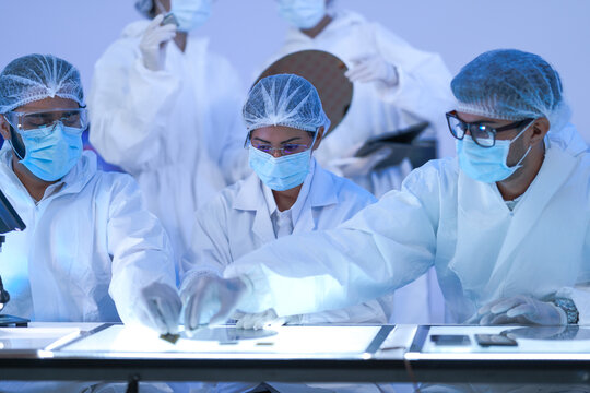 Two semiconductor engineers raise a wafer to the light for quality control in a microchip fabrication lab while wearing white lab coats and blue protective gloves.