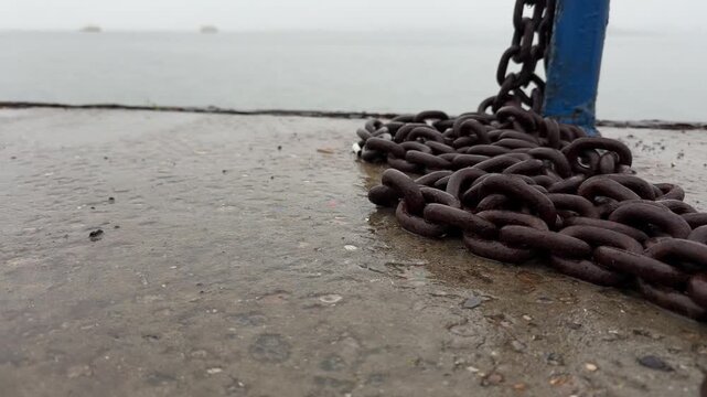 Rusty mooring chain piled on a wet concrete pier attached to a blue bollard, with two mooring dolphins visible in dense fog over calm river water in the background