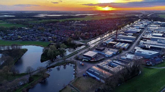 Aerial wide shot of a modern industrial zone and suburban residential neighborhood separated by a calm water canal in a European town at golden sunset