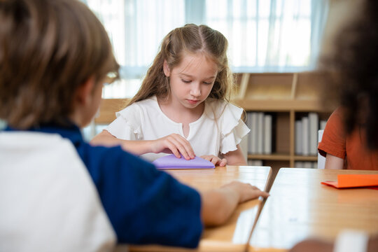 Elementary school girl concentrating on folding a purple origami paper project at her desk. Primary school student focusing on paper folding handicraft during arts and crafts lesson in classroom.
