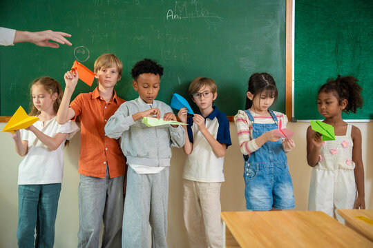 Diverse elementary school children standing in front of chalkboard holding colorful paper airplanes. Multiethnic primary students presenting their origami paper planes during creativity in classroom.