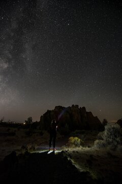 Young woman on a path shines torch, canyon against night sky, starry sky with Milky Way, Smith Rock State Park, Oregon, USA