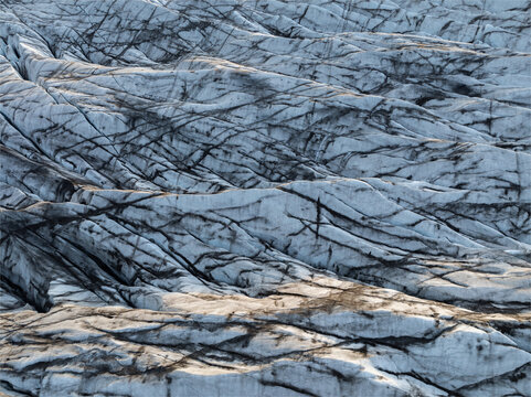 Aerial view of Svinafellsjokull glacier featuring intricate patterns of deep blue crevasses and dark volcanic ash deposits across the icy surface, Sveitarfelagid Hornafjordur, Iceland.