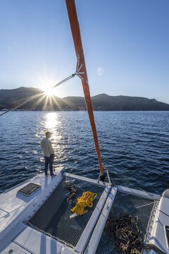 Young man standing on a catamaran looking into the sunset, ropes on a sailing catamaran, sun star, sailing trip, Tilos, Dodecanese, Greece