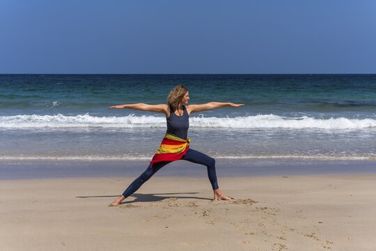 A woman doing yoga on a sandy beach by the sea. Virabhadrasana 2 or Warrior 2. Treen Cove, Cornwall, England, Great Britain