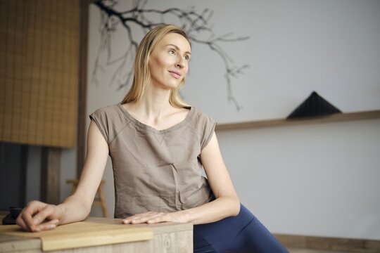 Cute woman sits at a wooden table in a simple room. She looks thoughtful while gazing in the distance. Natural light comes through the window, creating a warm atmosphere