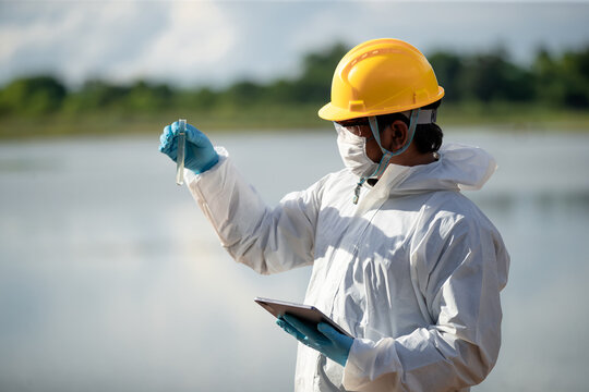 Ecologist examining water quality with flask and tablet near natural water source in protective gear