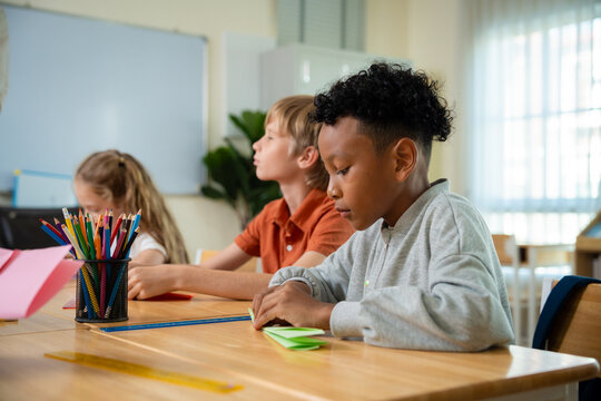 Young African American boy concentrating on folding paper airplane at a classroom desk. Primary school student making origami craft during creative arts and crafts lesson.