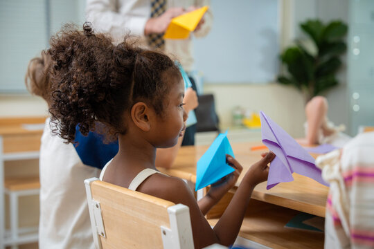 Cute schoolgirl holding colorful paper airplanes during an origami art class in classroom. Primary school child showing her handmade purple and blue paper planes with teacher in background.