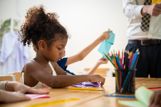 African American schoolgirl folding a purple origami paper at her desk during art class. Primary school children doing paper folding crafts with a male teacher in background.