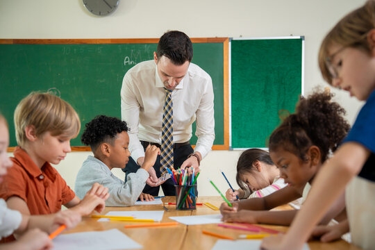 Male teacher guiding a diverse group of elementary school children during a drawing lesson. Professional schoolteacher supporting primary students writing and coloring at a classroom table.