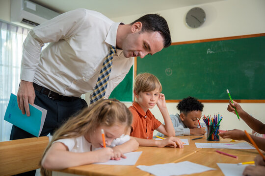 Male teacher bending down to help and talk to a primary school girl during a classroom lesson. Professional schoolteacher giving guidance and support to young students writing at a table.