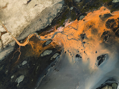 Aerial view of a glacial river delta with vibrant orange sediment flowing through dark volcanic sand channels in Iceland.