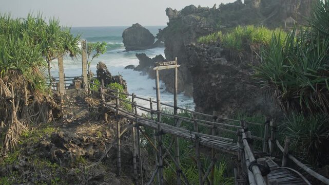 A rustic bamboo and wooden walkway stretches across a rocky tropical coastline toward crashing ocean waves, framed by pandanus plants and rugged cliffs for a secluded, peaceful seaside escape.