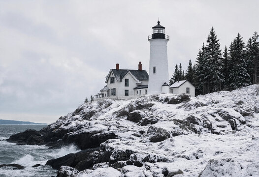 White lighthouse standing on a rugged rocky coastline covered in fresh winter snow