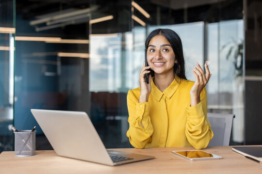 Indian smiling young businesswoman sitting in the office at a desk with a laptop and talking on the phone
