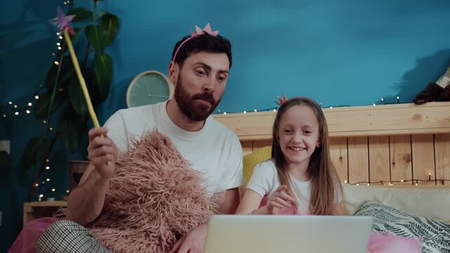 Fancy costume magic party at home. Portrait of happy dad and his lovable daughter waving with magic wand doing tricks watching videos on laptop in the bedroom.