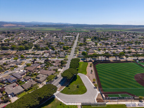 Aerial view showing a baseball field with striped turf next to a residential neighborhood, with streets, houses, and distant mountains under a clear sky in Salinas, California