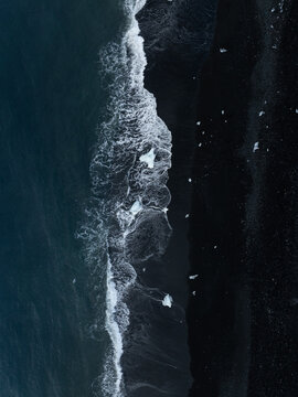 Aerial view of Diamond Beach with scattered ice chunks on the black sand shore and crashing ocean waves in Sveitarfelagid Hornaf, Iceland.