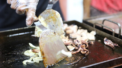Street food vendor cooking grilled squid octopus seafood on hot black iron griddle plate using metal spatula with plastic gloves at Asian night market. © Radhitya Ardian Arka