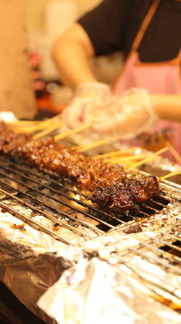 Close up of delicious grilled honey pork satay sate babi madu skewers cooking on charcoal grill with smoke, vendor wearing pink apron and plastic gloves handling satay.